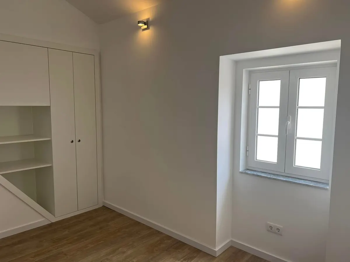 Depois da obra: quarto com teto inclinado, roupeiro embutido, aplique de parede e janela nova.